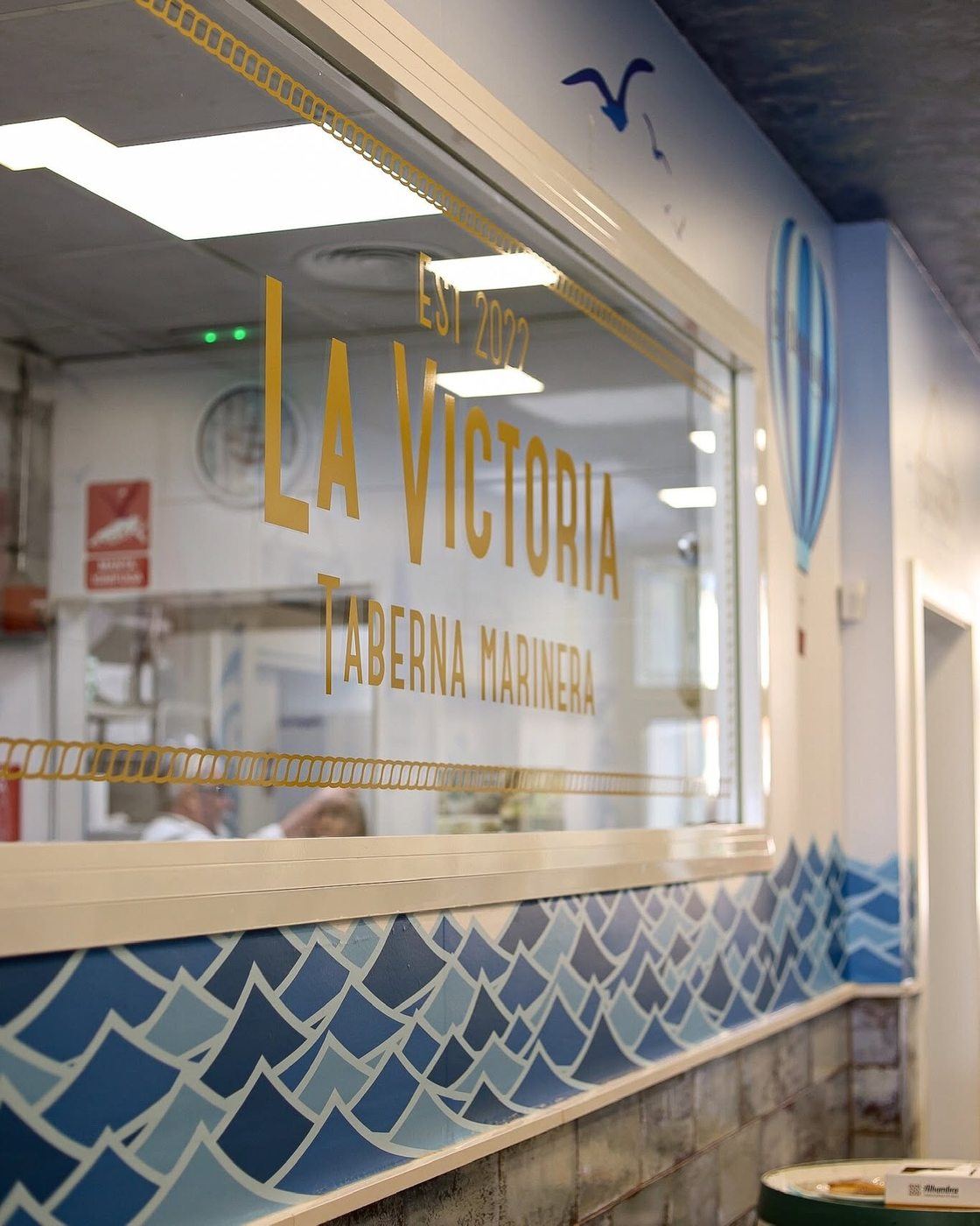 Ventana con el rótulo dorado de La Victoria, Taberna Marinera, junto al azulejo de olas
